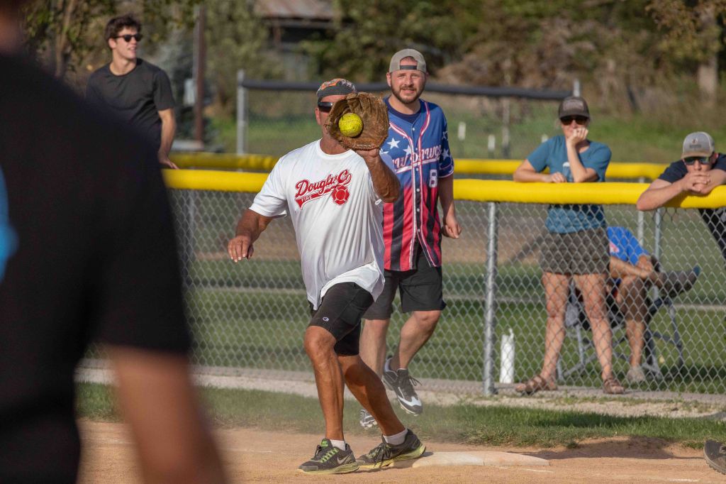 A player in a white shirt throws a softball while standing on a baseball field, with spectators in the background watching the game.