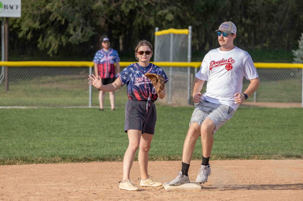 A young woman in an American flag-themed shirt plays softball, with a baseball glove in hand, showing a defensive stance. A man in a white shirt and shorts runs towards the base, while another person in the background observes the game.