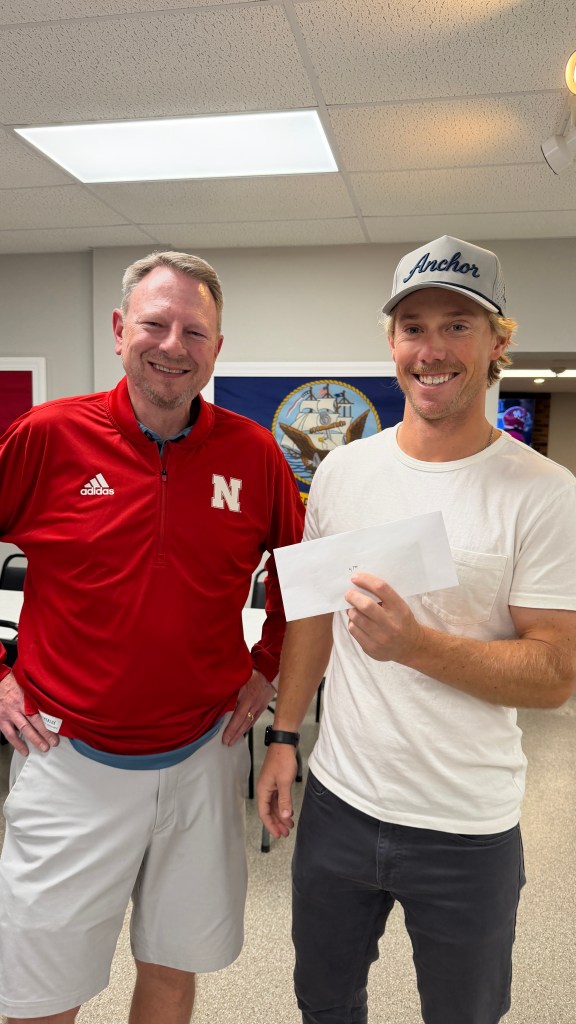 Two men are standing together in an indoor setting. The man on the left is wearing a red athletic jacket with a Nebraska logo, while the man on the right is holding an envelope and wearing a gray cap with 'Anchor' written on it. They are smiling and appear to be having a friendly conversation.