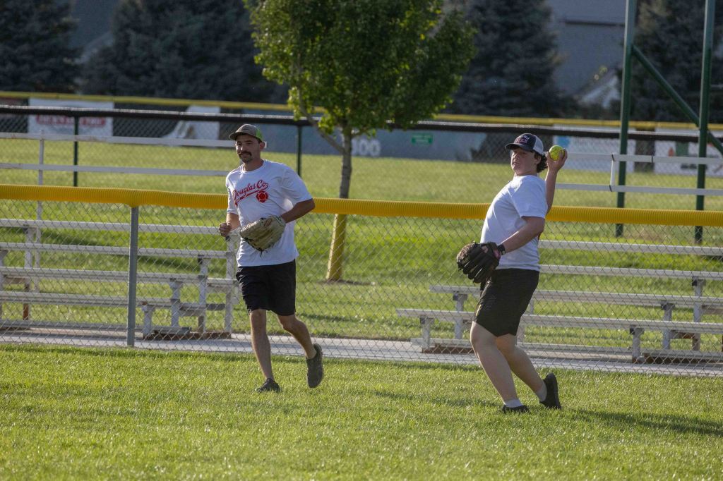 Two men playing catch on a softball field, one running with a glove and the other preparing to throw a ball.