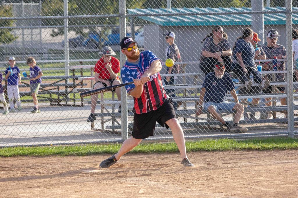 A player in an American flag-themed shirt swings a bat at a softball during a game. Spectators watch from the bleachers in the background.