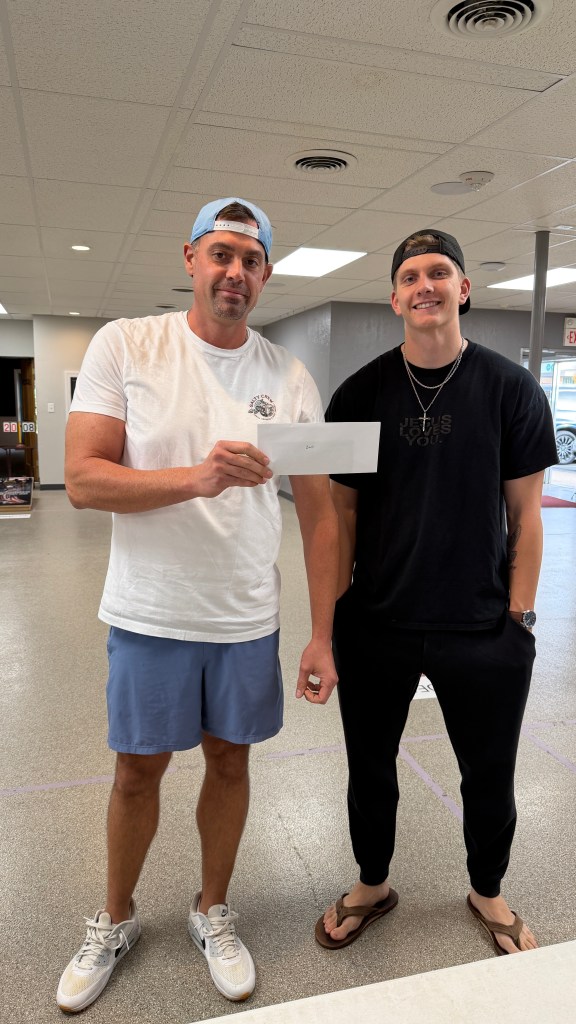 Two men standing indoors, one holding an envelope. The man on the left is wearing a white t-shirt and blue shorts, and the man on the right is in a black t-shirt and pants, smiling.