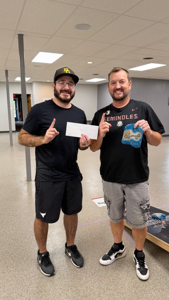 Two men standing together in an indoor space, one holding a check and the other holding a cookie package, both smiling and appearing happy.
