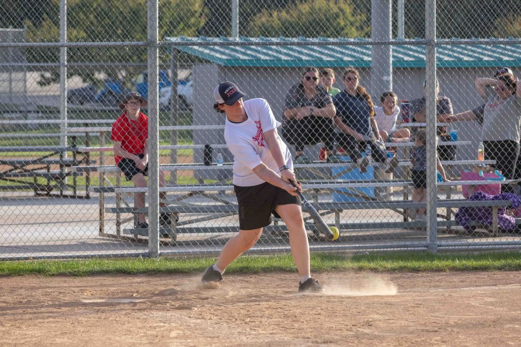 A young person swings a bat while playing softball in a dusty field, with spectators in the background watching the game.