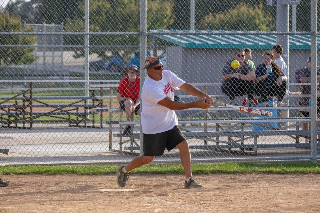 An adult man swings a softball bat during a game at a local park, with spectators sitting on bleachers in the background.