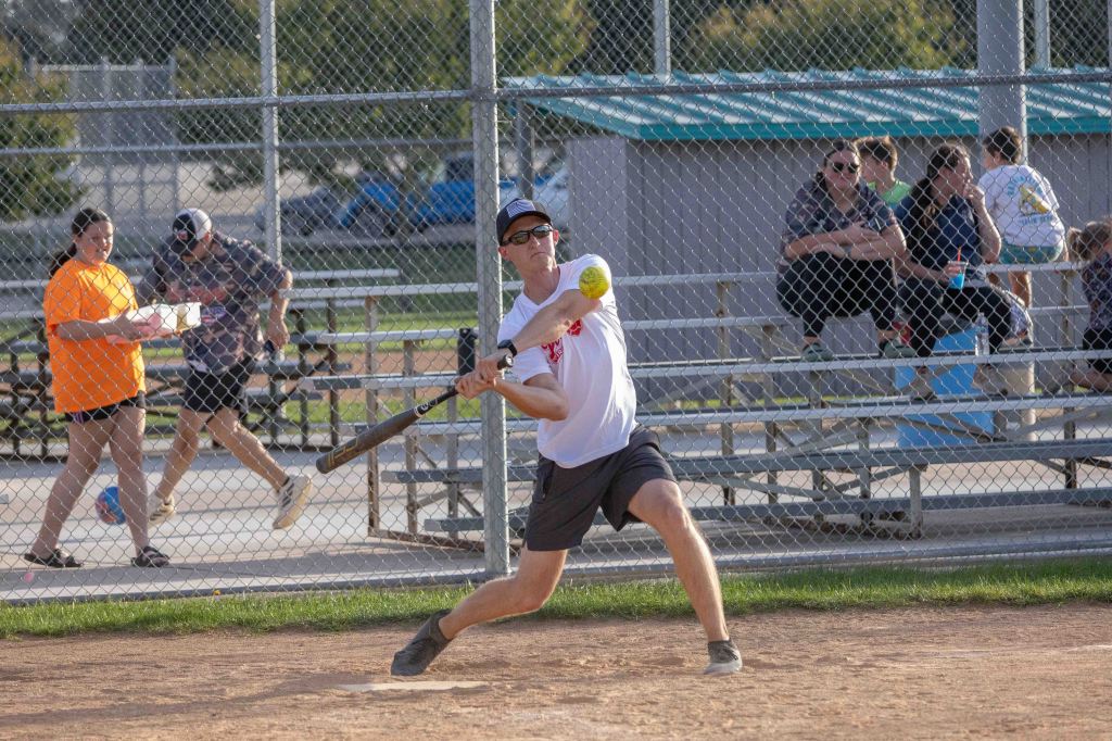 A young man swings a bat to hit a softball during a game at a park, while spectators watch from the bleachers.