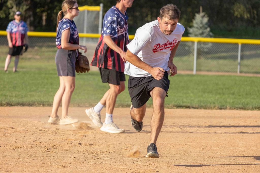 A male player running on a softball field, wearing a white shirt and black shorts. In the background, a female player in a navy top with red sleeves and other players can be seen.