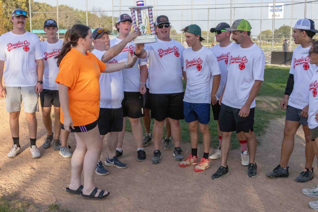A group of young men wearing matching white T-shirts with 'Douglas Co' printed on them stand together at a softball field, while a woman in an orange shirt is holding up a trophy.