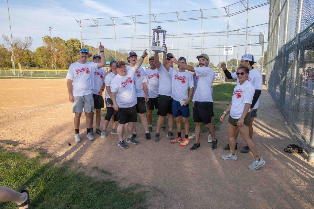 A group of eleven people wearing matching white shirts posed together on a baseball field, celebrating with a trophy raised high. They are showing excitement with their arms raised and smiling.
