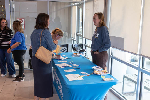 A group of people interacting at a registration table in a bright, open space. One woman in a blue shirt is speaking with another at the table, which is covered with a blue cloth and has snacks and informational materials on it.