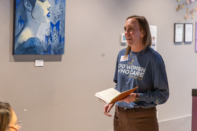 A woman standing while speaking to an audience, wearing a blue shirt with the text 'DO WOMEN WHO CARE'. She holds a notebook in her hand and is positioned near an abstract painting on the wall.
