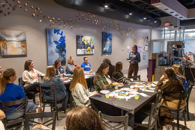 A group of women seated at a table participating in a creative workshop, with a presenter speaking in front of them. The room has colorful artwork on the walls and a relaxed atmosphere.