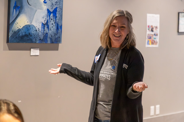 A woman with shoulder-length hair smiles and gestures while standing in front of a blue-themed artwork on the wall.
