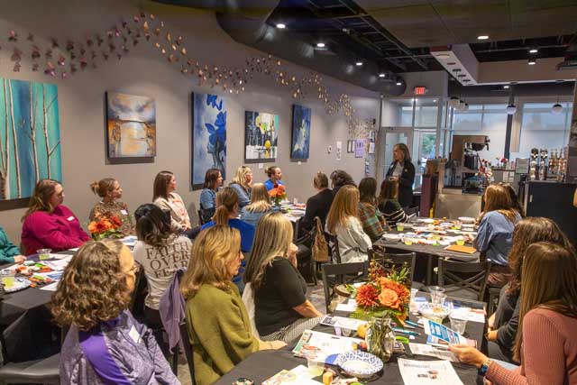 A group of women attending a workshop or seminar in a cozy indoor setting, sitting around tables decorated with flower centerpieces and various materials. An instructor stands at the front, engaging with the audience, while colorful artwork adorns the walls.
