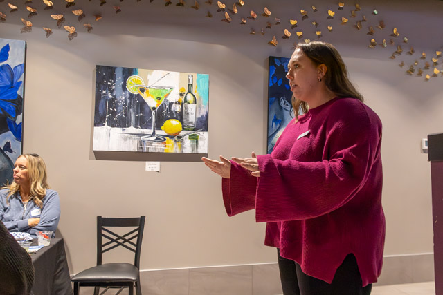 A woman in a maroon sweater is speaking while gesturing. In the background, there are colorful paintings of cocktails and flowers. A seated woman is visible on the left.