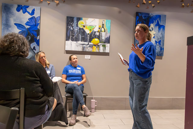 A woman in a blue sweater passionately speaks to a small group seated in a cozy setting, with colorful artwork on the walls.