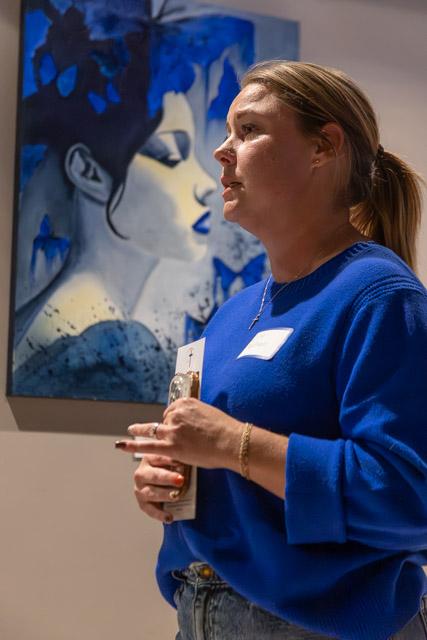 A woman in a blue sweater speaks while holding a phone, with a large blue and gray painting of a woman's face in the background.