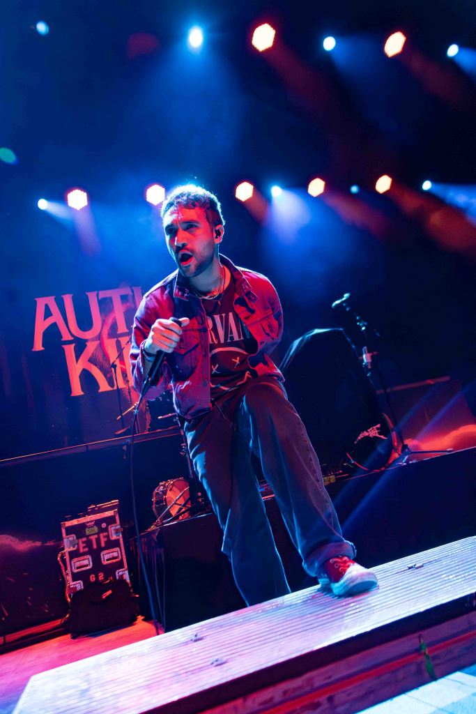 A male singer performing on stage with intense energy, wearing a graphic t-shirt and a denim jacket, surrounded by colorful stage lights.