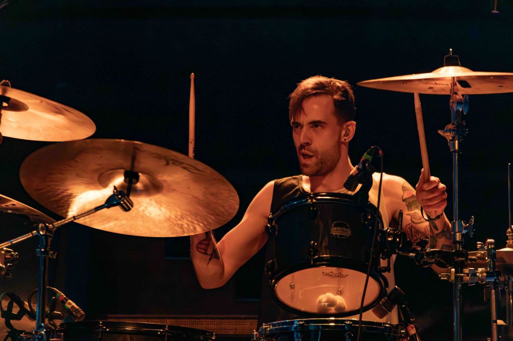 A drummer performing on stage with drumsticks raised, focused on playing a drum set under soft stage lights.