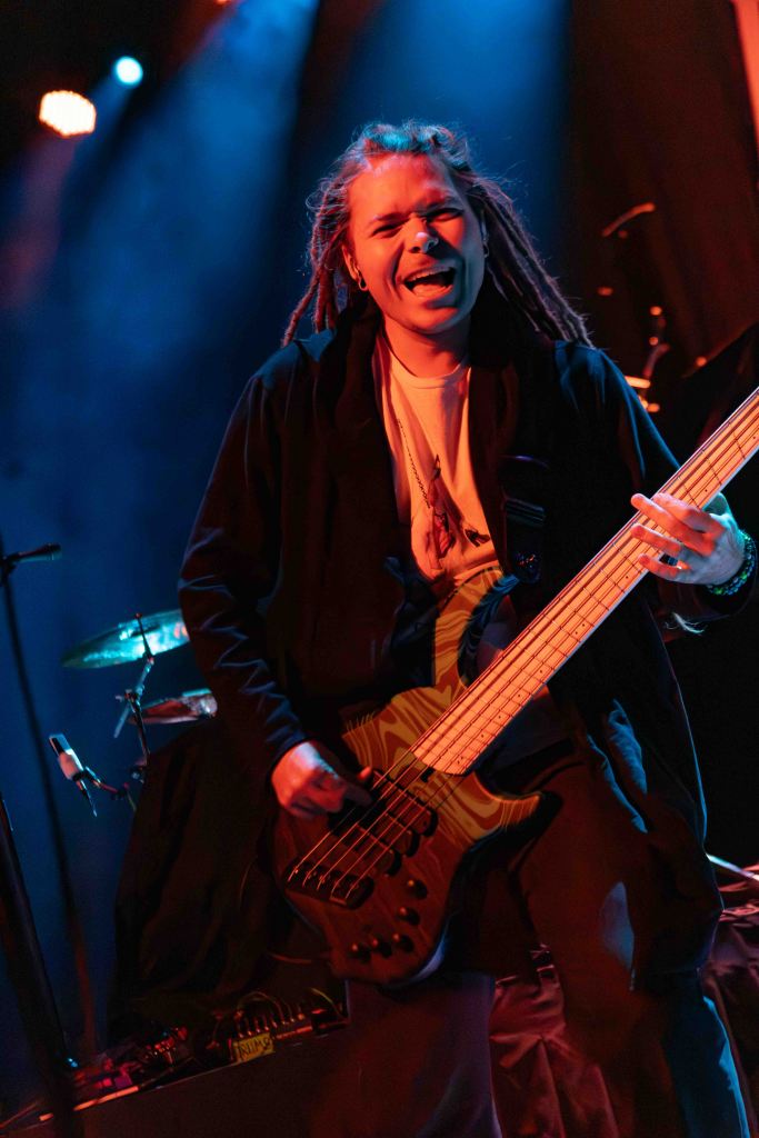 Musician with dreadlocks passionately playing an electric bass guitar on stage under blue lighting.