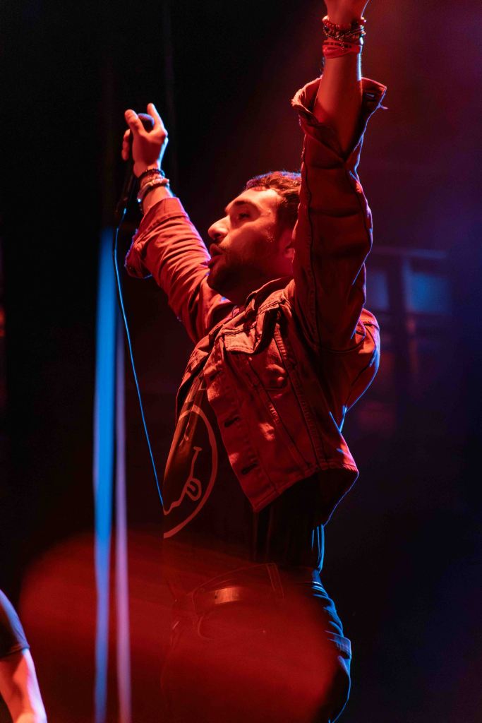 A performer on stage raising both hands in the air, wearing a denim jacket and black shirt, with dramatic lighting creating a vibrant atmosphere.