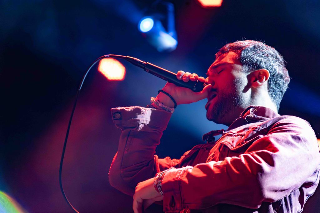 A male singer passionately performing on stage with a microphone, under colorful lighting.