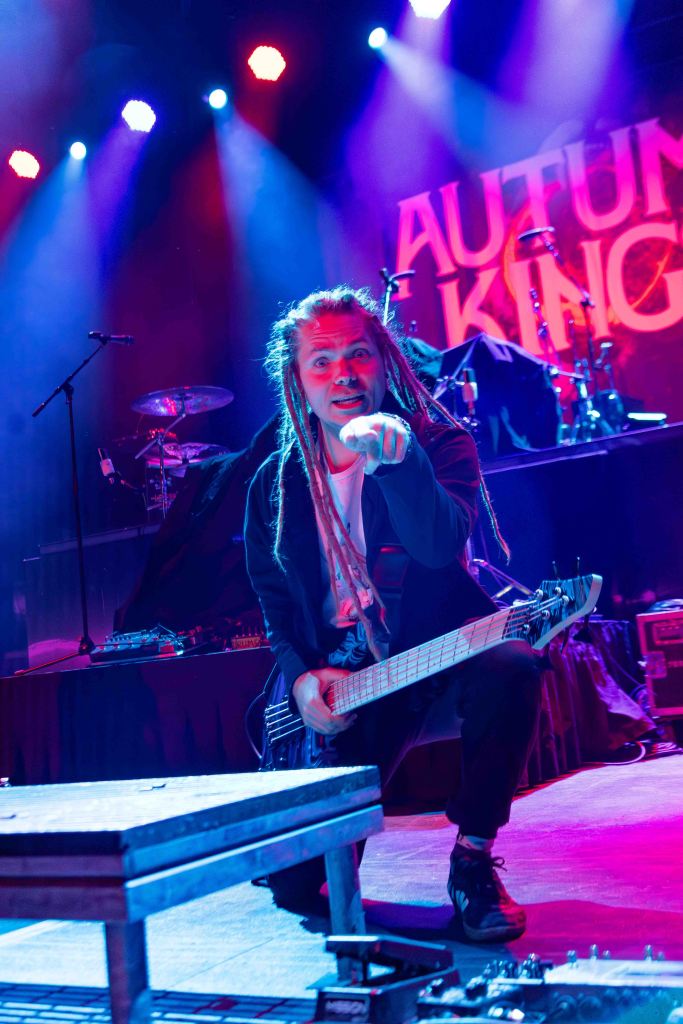 A musician with long hair and dreadlocks squats on stage, holding a bass guitar and pointing towards the audience. Colorful stage lights illuminate the scene, with a backdrop displaying 'AUTUMN KING.'
