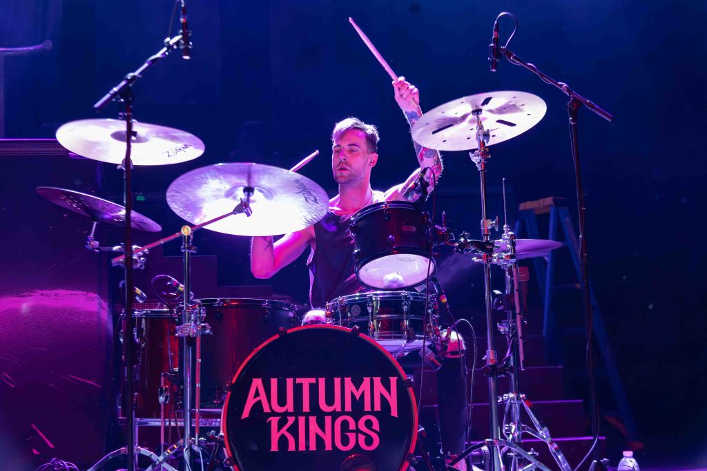 A drummer performing on stage with a drum kit and cymbals, featuring the name 'Autumn Kings' on the bass drum.