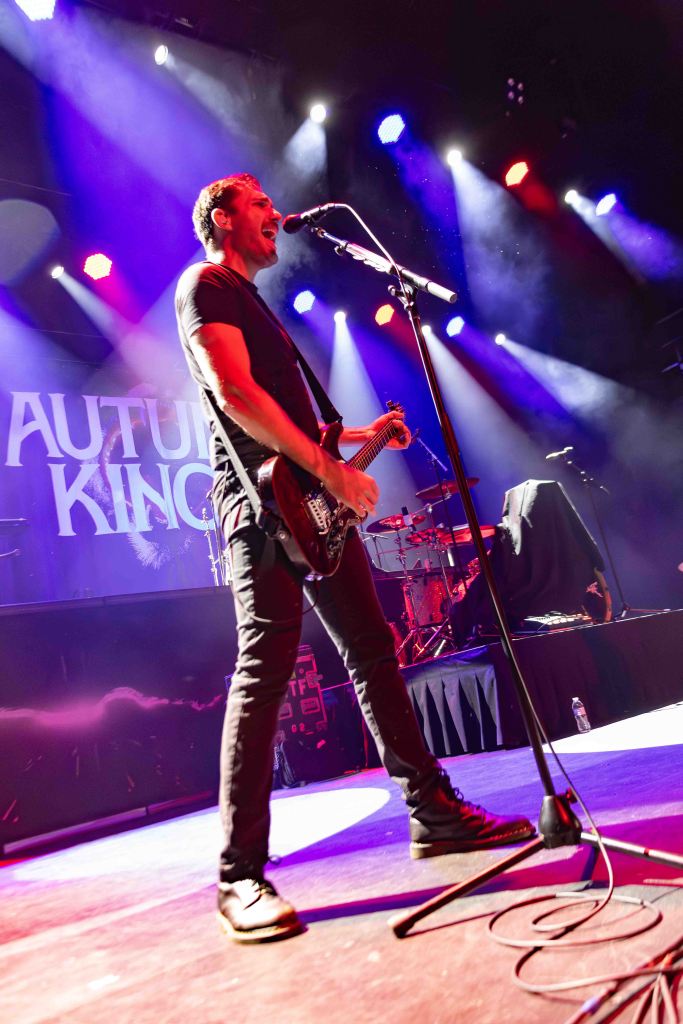 A musician performing live on stage, singing into a microphone while playing an electric guitar. Colorful stage lights illuminate the scene, creating a vibrant atmosphere.