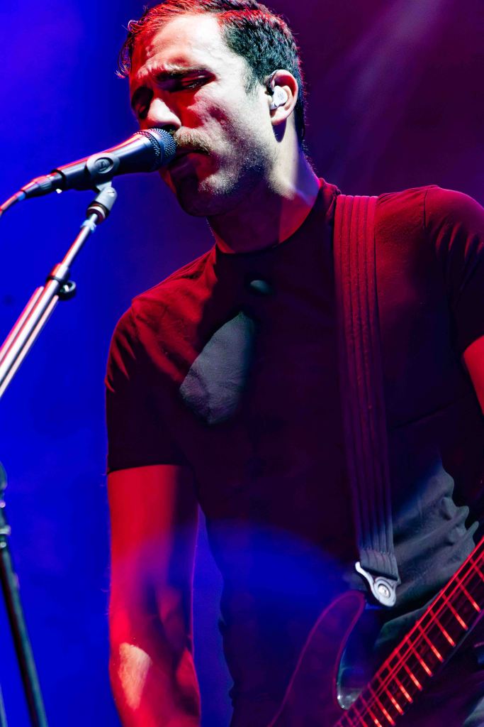 A male musician singing into a microphone while playing an electric guitar, illuminated by colorful stage lights.