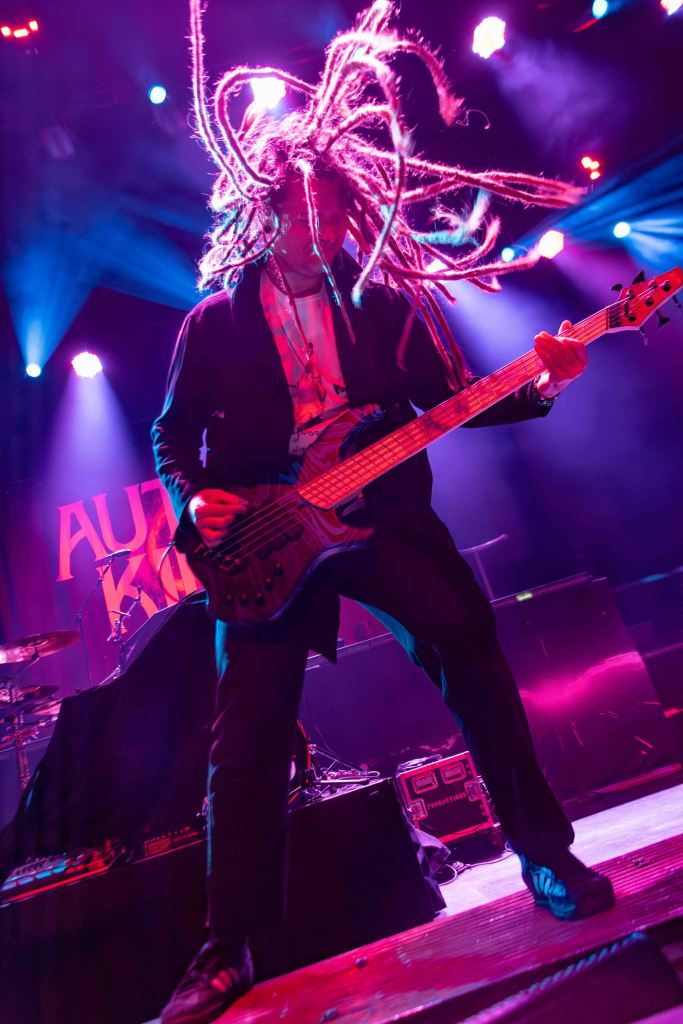 A musician with long dreadlocks passionately playing a bass guitar on stage, surrounded by colorful stage lights.