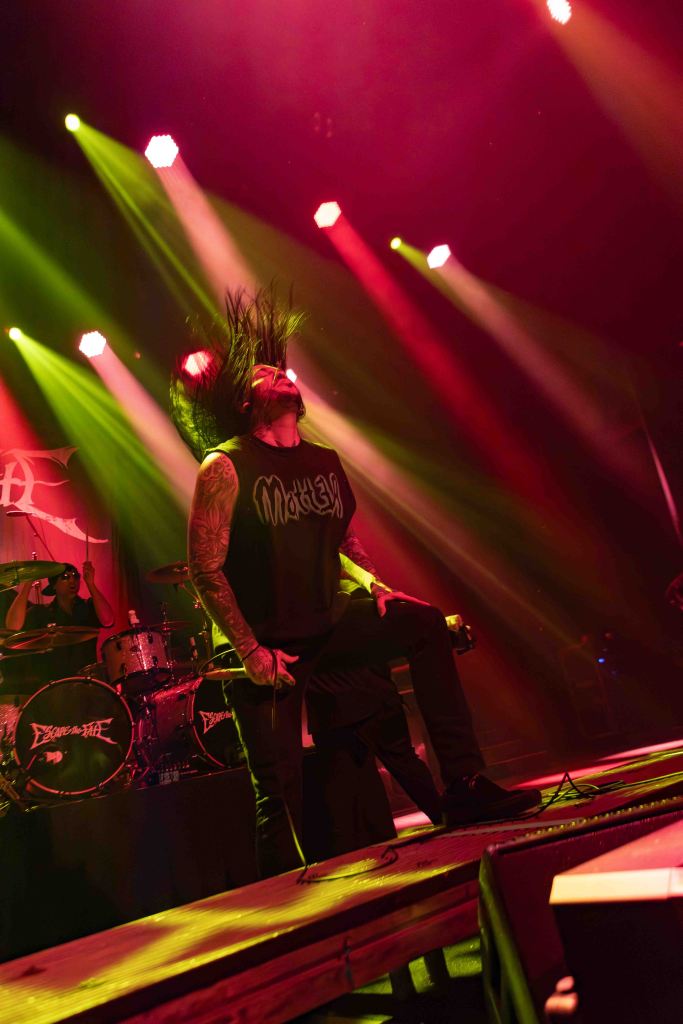 A musician performing on stage with long hair moving energetically under colorful stage lights, wearing a black sleeveless shirt with tattooed arms. A drum set is visible in the background.