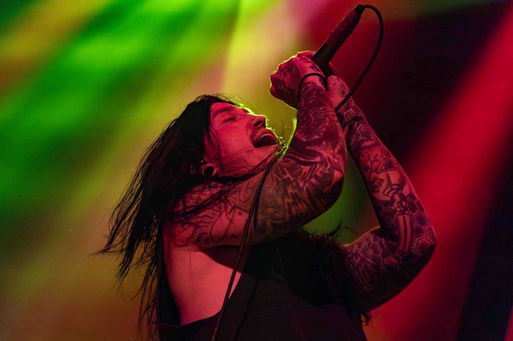 A singer with long hair and extensive tattoos performs passionately on stage, holding a microphone with intense expression, illuminated by colorful stage lights.