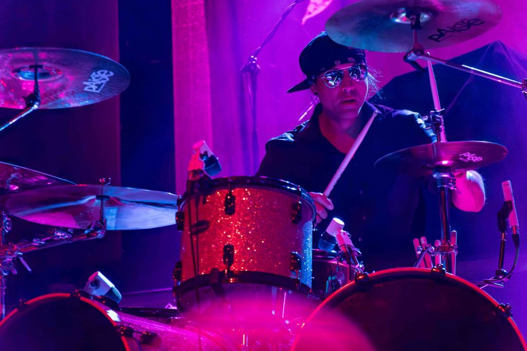 A male drummer performing on stage, wearing sunglasses and a black cap, surrounded by colorful stage lighting.