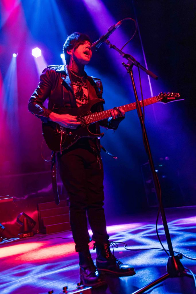 Musician performing on stage with a guitar, wearing a black leather jacket, under colorful stage lights.
