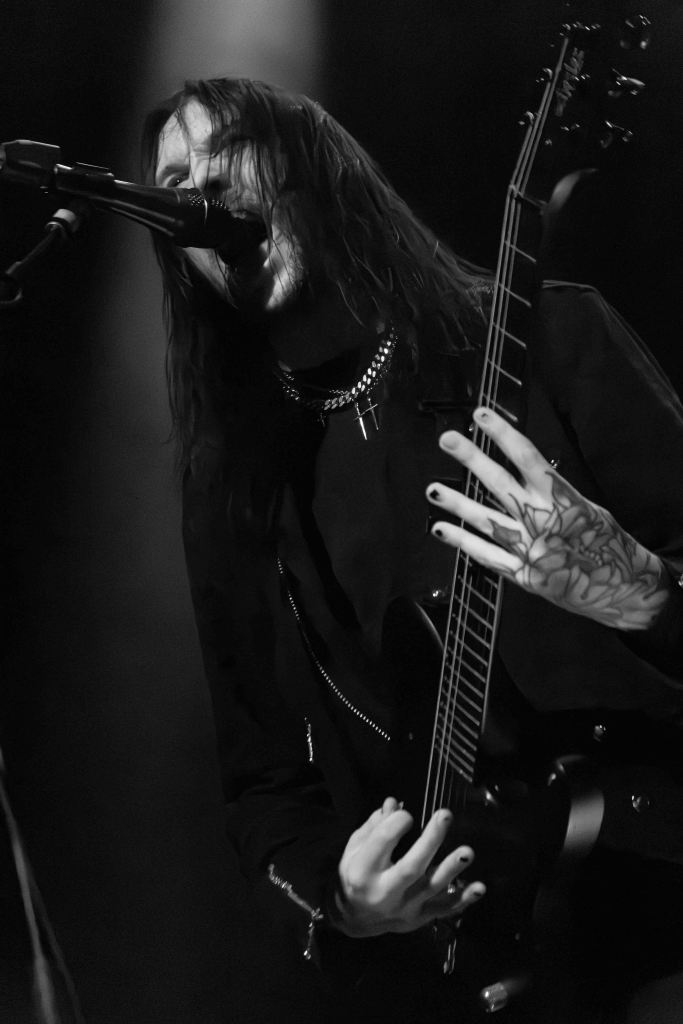 A musician performing on stage, passionately singing into a microphone while playing an electric guitar. The image is in black and white, highlighting the intensity of the performance.