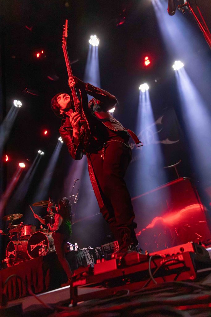 A guitarist performing on stage with dramatic lighting and a drummer in the background, creating an energetic atmosphere.