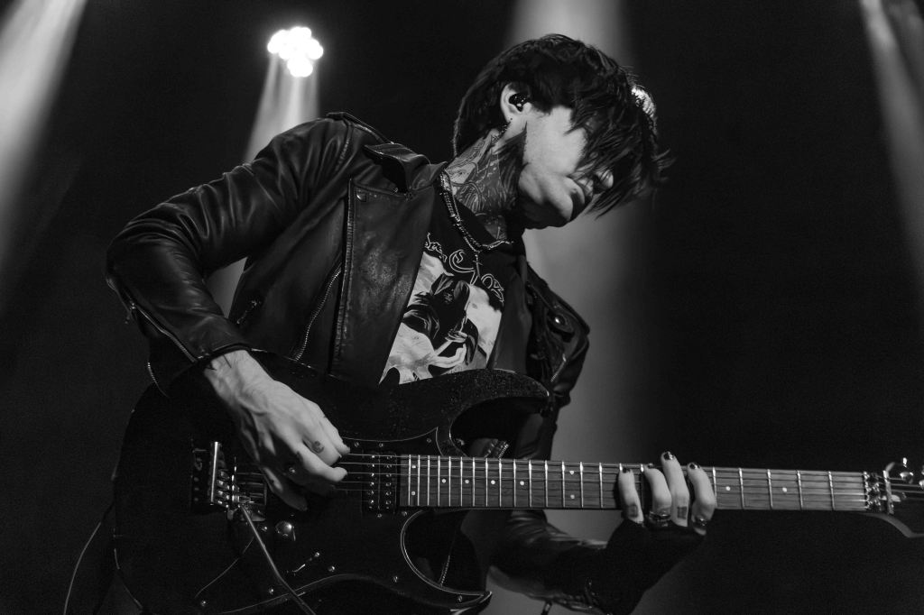 A musician wearing a leather jacket passionately playing an electric guitar on stage, captured in black and white.