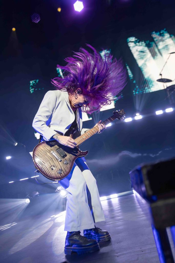 A musician performing on stage with vibrant purple hair, wearing a white suit, passionately playing an electric guitar under dynamic stage lighting.
