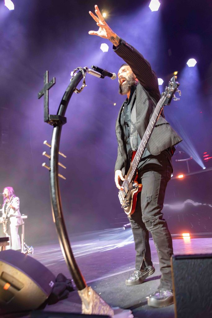 A male bassist performing on stage, passionately raising one hand while playing his instrument. The background features colored stage lights and another band member in the distance.