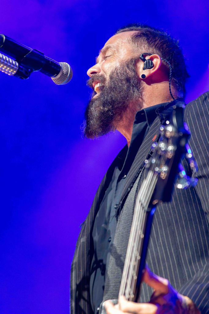 Close-up of a musician with a beard singing into a microphone while playing a bass guitar, illuminated by colorful stage lights.