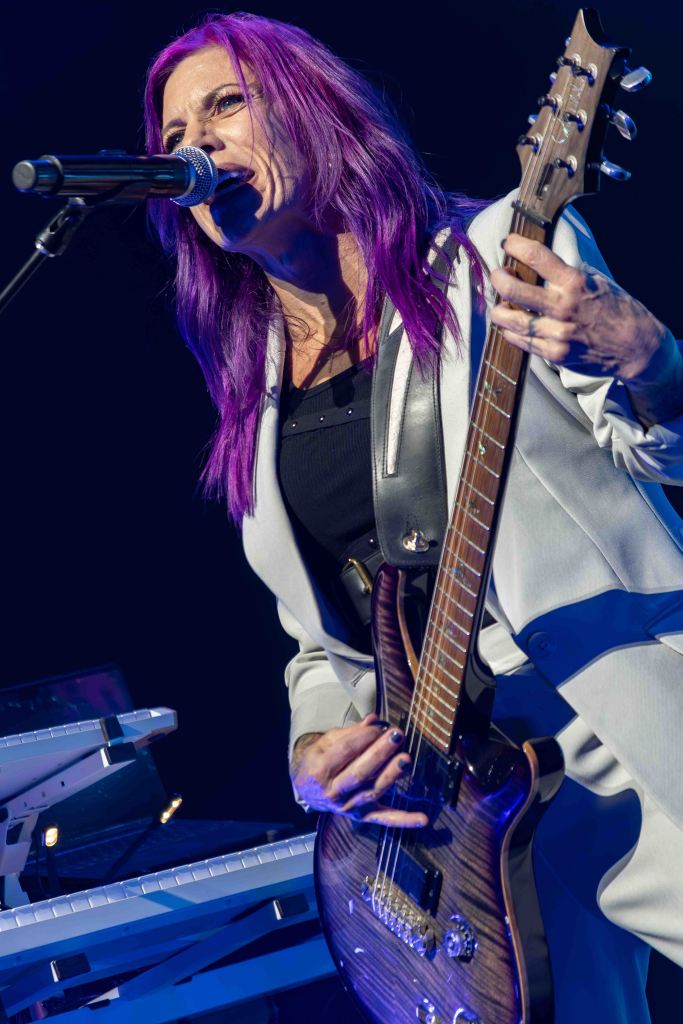A female musician with long purple hair performing on stage, singing into a microphone while playing an electric guitar. She is wearing a stylish outfit, with a keyboard visible in the background.