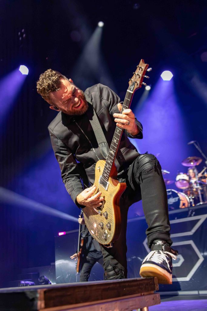 A guitarist passionately performing on stage with dramatic lighting and a lively concert atmosphere.