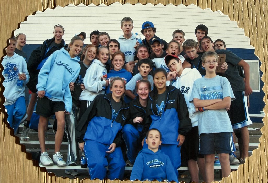 A large group of high school students wearing matching blue and black athletic gear, posing together and smiling for the camera, with a school building in the background.