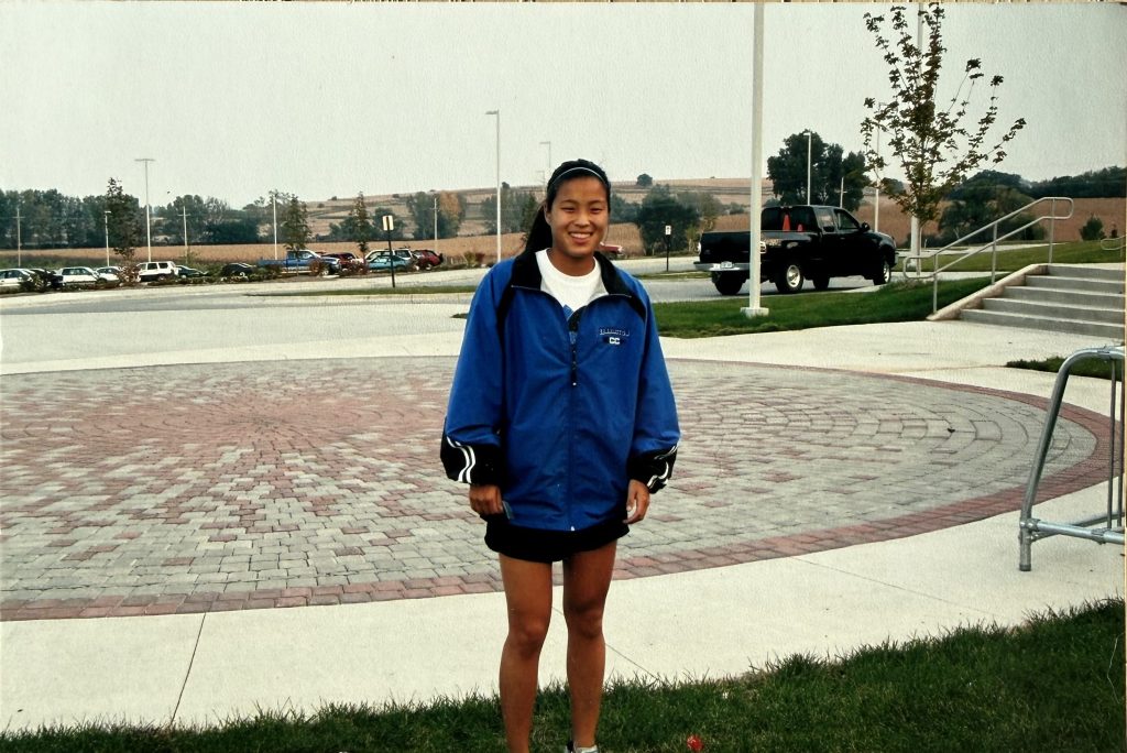 A young athlete stands outdoors in a blue jacket and black shorts, smiling in front of a paved area with trees and cars in the background.