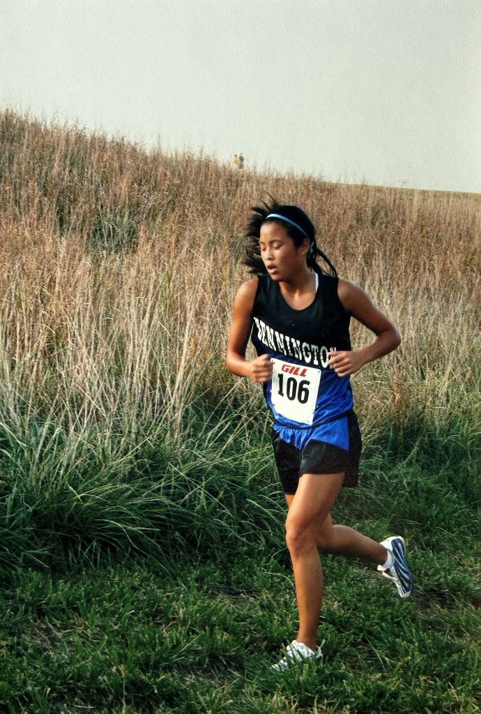 A young female runner wearing a black tank top and blue shorts, featuring a race number 106, running through a grassy area with tall grass in the background.