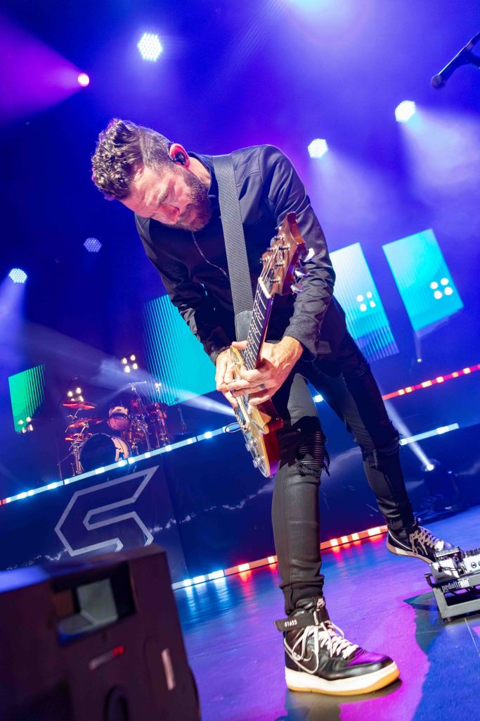 A guitarist performing on stage, bending forward while playing an electric guitar. The background features colorful stage lights and a drummer is visible behind the guitarist.