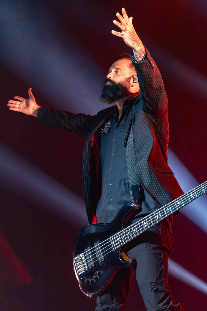 A male musician performs on stage with outstretched arms, holding a black bass guitar, dressed in a pinstriped blazer and shirt, with dramatic lighting in the background.
