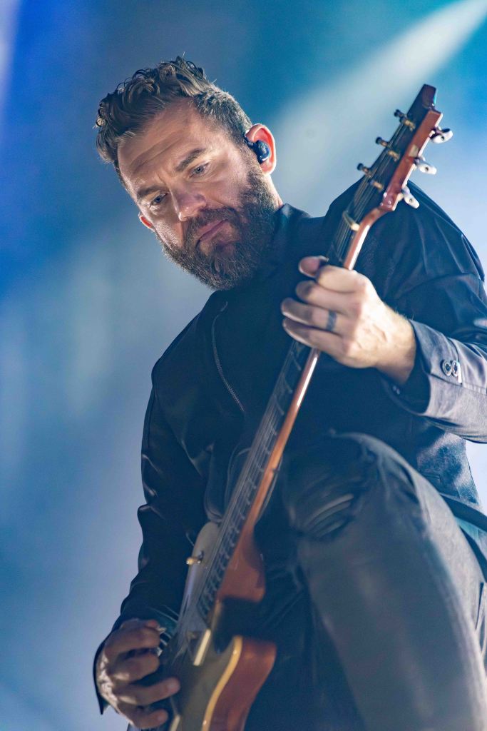 A musician with a beard performing on stage, playing an electric guitar. The background is illuminated with soft blue lighting, creating an atmospheric effect.