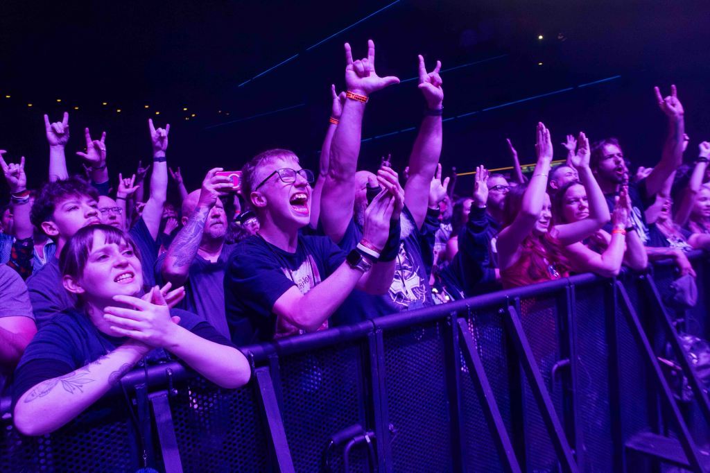 A lively crowd at a concert, with fans cheering and waving their arms, some making rock hand gestures, set against a purple-lit background.